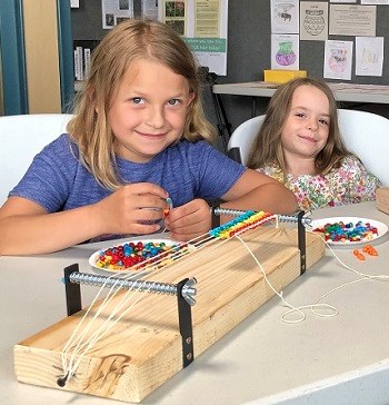 2 girls sit next to a bead loom indoors and smile. 1 girl puts beads on a needle.