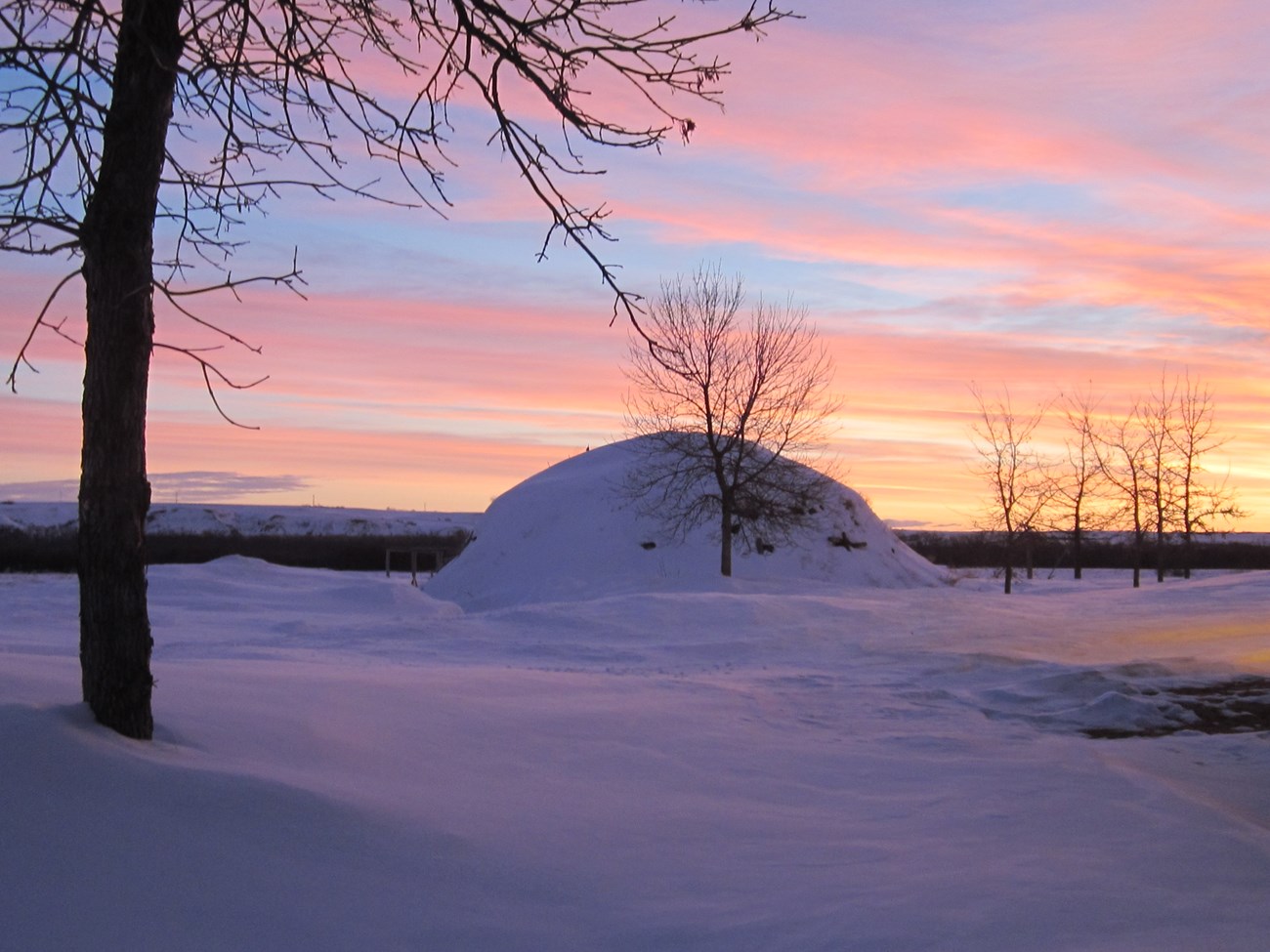 An earthlodge covered in snow with a colorful sky above.