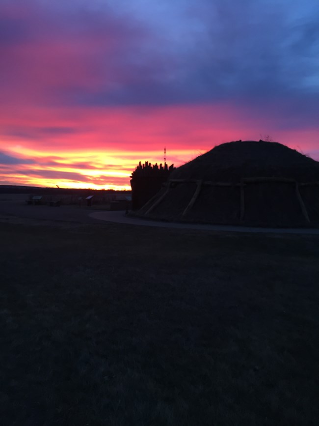 An earthlodge's silhouette against a colorful sunrise.