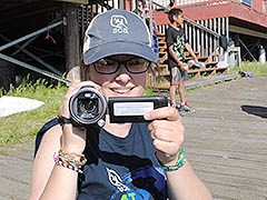 Young woman holding a video camera standing on a boardwalk. Child stands in background.