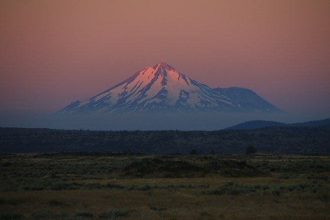 A triangular snow-capped mountain at sunrise, with low clouds around the base and rolling green land in the foreground