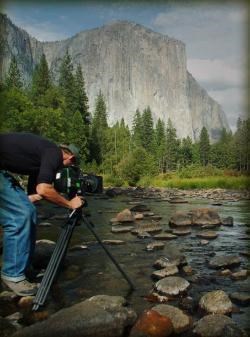 A photographer operates a camera and tripod near a stream and granite mountains at Yosemite National Park