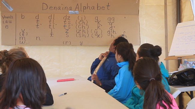 Students sit at table with cardboard sign reading "Dena'ina Alphabet"
