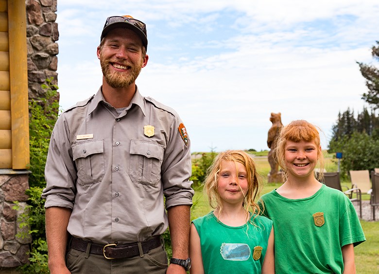 Two children wearing green shirts and a junior ranger badge stand next to a park ranger outside on a lawn.