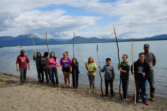 Middle School and High School age students, a park ranger, and an archeologist stand by a lake with mountains in the background.  Students have long spears in their hands.
