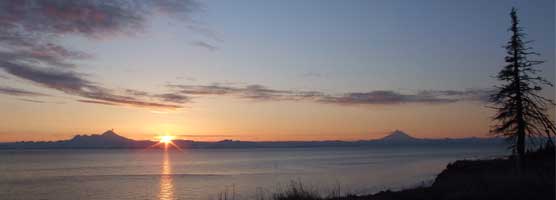 Silhouettes of steaming Iliamna and Redoubt Volcanoes viewed at sunset across Cook Inlet. Sun setting between the volcanoes. It's light reflected on the water.