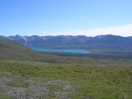 Green tundra hills in foreground extending down to a small sliver of turquoise colored lake backed by mountains.