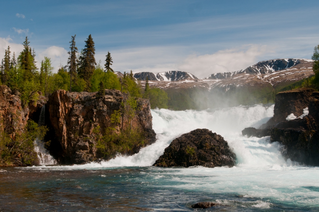 White frothy water cascading around large boulder surrounded by forested cliffs. Mountains in the background.