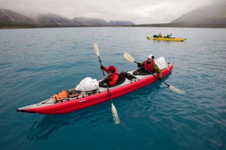 Red double hole kayak on turquoise-colored lake with foggy mountains in the distance. Warmly dressed young person paddling with double bladed paddle in front, wamly dressed adult paddling in rear. Yellow kayak in distance.
