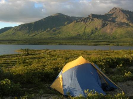 Blue and yellow dome tent catching evening light surrounded by low green vegetaion. Blue lake, green mountains and grey clouds in the distance.