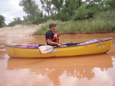 A paddler in a yellow canoe is headed down the reddish-orange Canadian River.  There are green cottonwoods in the background.  The paddler has on a blue shirt and hat.