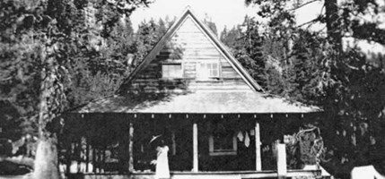 black and white photo showing rustic cabin in the woods with lady and umbrella on porch