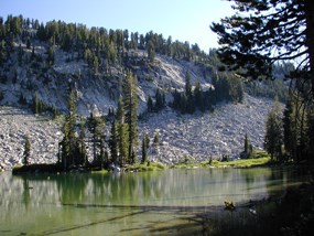 view of emerald green cliff lake with trees reflecting on calm water and rocky reading peak in the background.