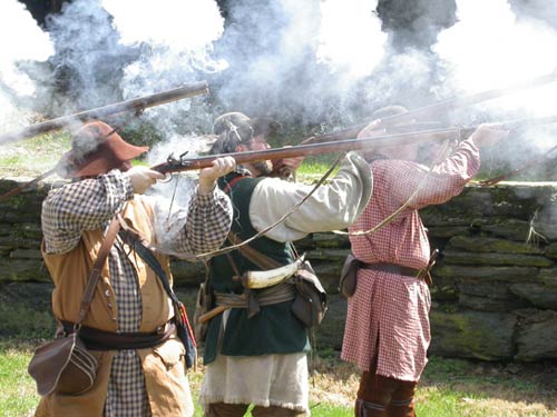 Reenactors dressed in period clothing fire rifles as gun smoke hangs in the air.