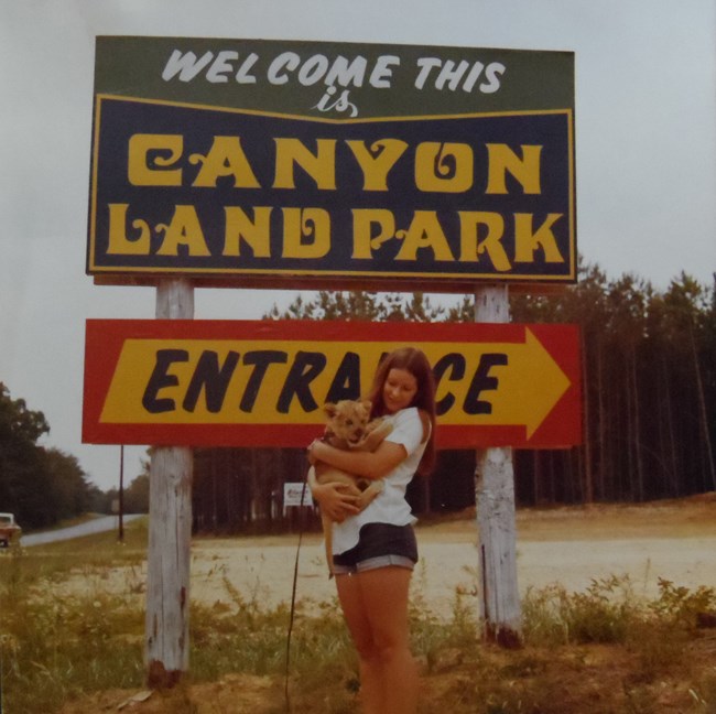 A young lady holding a lion cub stands beneath the entrance sign for Canyon Mouth Park & Zoo.