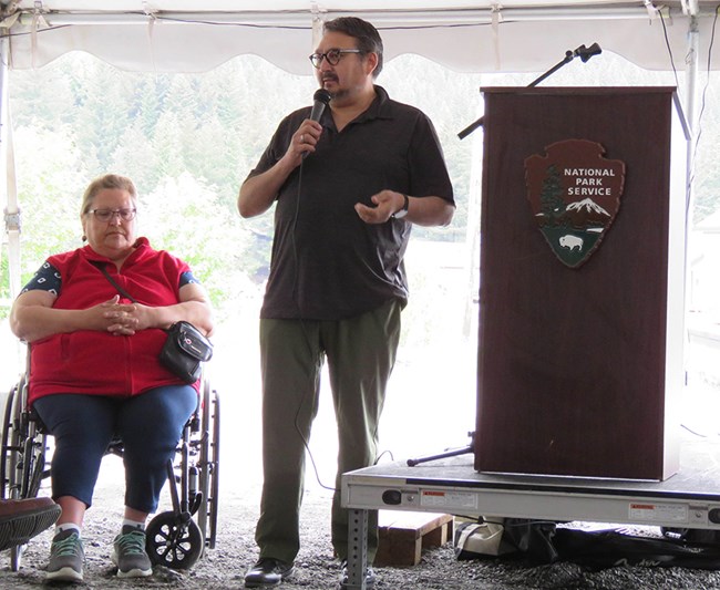 A woman in a wheelchair and a man on a stage.  They are next to a podium with the National Park Service Logo.