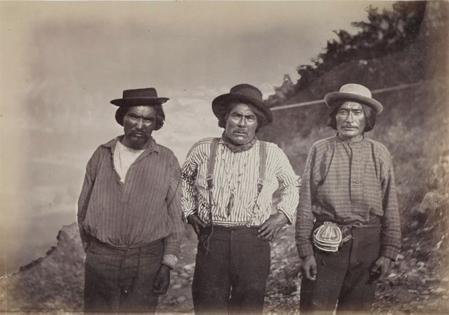 Three Alaska Native men dressed in hats and button-down shirts stand beside a river.