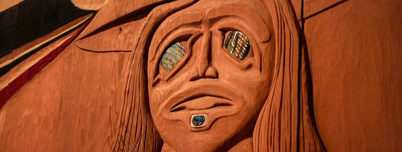 a face with long hair is carved into wood with shiny eyes.