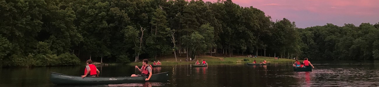 A group paddles canoes under a pink sky at dusk.