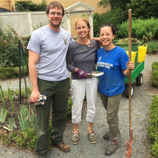 National Park Service staff member with two volunteers in the historic garden, holding gardening tools