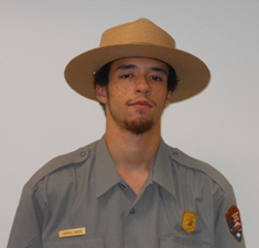 A young male park ranger stands against a grey wall to have his picture taken