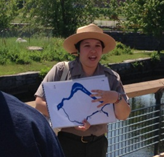 A young female park ranger holding a map while standing in front of a canal and talking to visitors.
