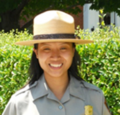 A young woman, dressed as a park ranger, stands in front of some shrubs and smiles
