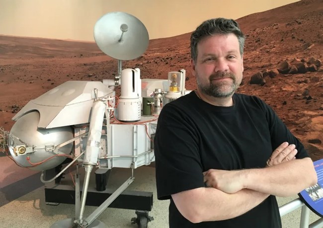 A man stands in front of a space vehicle model with a Mars photo in the background.