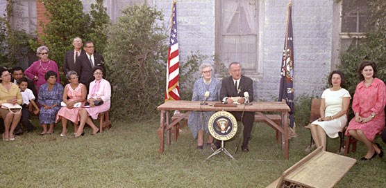 President Lyndon B. Johnson addresses crowd while Lady Bird Johnson and Lynda Bird Johnson listen. Seated next to the President is Kate Deadrich Loney. 