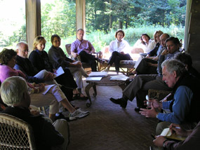A group of 13 people sit in earnest conversation on the wicker chairs of a sunlit porch, vibrant green woods behind them.