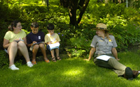 Three children and a park ranger sit in the shady grass and work on Junior Ranger actitivies.