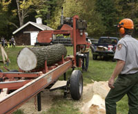 A park ranger in an orange hardhat and orange ear protectors watches as a portable saw mills a thick log.