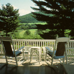 Two white wooden and wicker rocking chairs sit on the Mansion porch, looking out over fields and hills. Photo by Nora Mitchell.