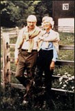 This color photograph shows Laurance Rockefeller on the left and his wife Mary on the right, standing in front of a split rail wooden fence and a barn, with white flowers peeking through the rails. They both have sweaters tied around their necks.