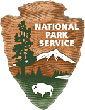 The symbol of the National Park Service, the brown chiseled arrowhead logo depicts a bison, an evergreen tree, a lake, and a snow-covered mountain peak.