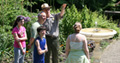A family in brightly-colored clothing speaks with a ranger around a garden fountain; the ranger is pointing to something in the distance. NPS Photo.