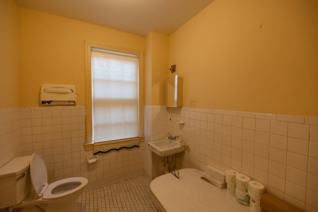 A toilet sits on the left. Directly across is a sink and mirror. In the foreground is a covered, non-functioning bathtub.