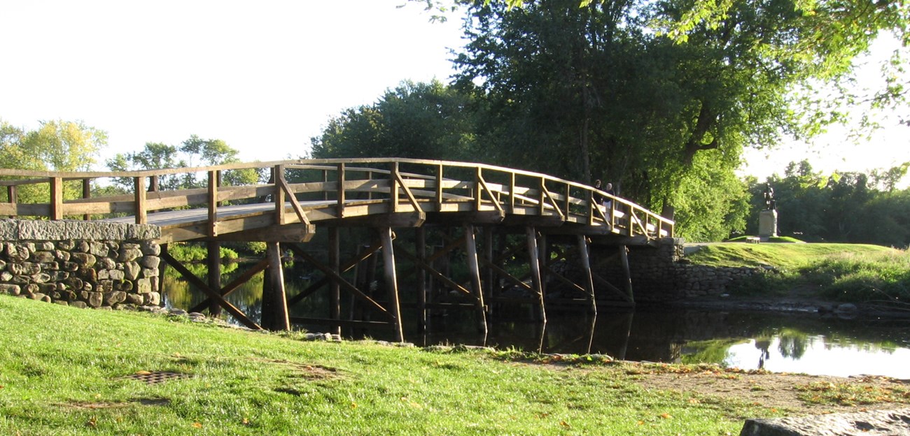 A slightly arched wooden bridge spans the Concord river. Green grass covers the river banks and tall trees rise from the opposite bank.