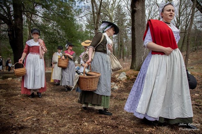 Women in colonial clothing bearning baskets and cloth bags walk through a wooded landscape