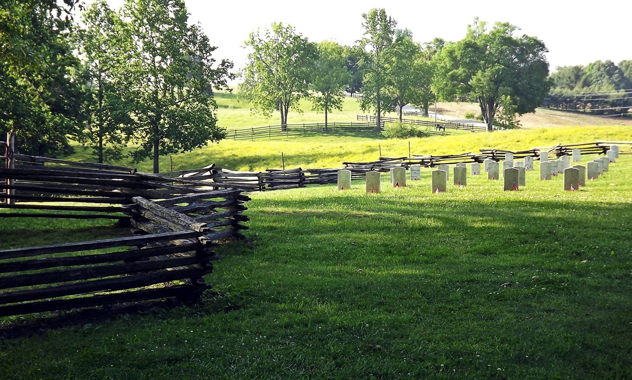 Headstones and split rail fence in rolling grassy field with roadway passing through in distance