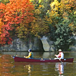 Mississippi River canoeists glide past trees with red and gold leaves perched on a riverside cliff.