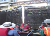Canoes locking through a dam. Concrete walls loom on either side and the control building sits high above.