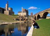 A person sits at the edge of a canal watching a Canada goose. Old mill district with ruins are in the background.