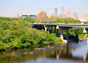 Canoeing the Mississippi River. A large bridge and city skyline rises behind the canoes.