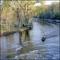 A late afternoon sun illuminates the wooded river banks. A motor boat runs down the Rum River.