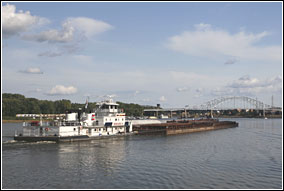 A large, white tow boat pushes a raft of barges downstream on the Mississippi River under blue sky and white, puffy clouds. A large bridge is in the distance.
