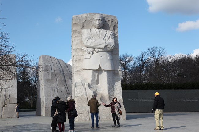 A tall cream-colored statue of Martin Luther King, Jr. stands in an open area. In front of the statue, visitors cross and chat.