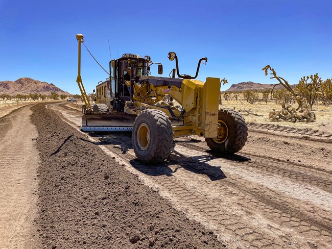 A yellow road grader with GPS receiver antenas on the blade travels along a dirt road