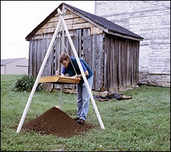 A worker sifts dirt through a screen hanging from a tripod. A small wood building in the background.