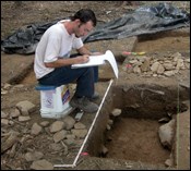 An archeologist writes notes while sitting next to a rectangular hole.